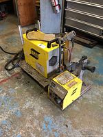 Front view of yellow ESAB PCM-1125 power supply unit with handles, connected cables, Mig 4HD feeder on right, pair of yellow leather gloves on top