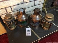 Three vintage kerosene lamps with one labeled 'Standard', one 'Port', and one 'Starboard' shown together on dark surface in front of stone wall.