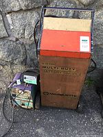 Wide photo showing three battery chargers: Marquette Multi Duty Battery Service with large orange and beige wheeled unit, and two smaller vintage chargers with visible rust and wear.