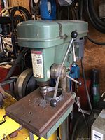 Front and side view of green Olympic GL 13 drill press showing main body, work table, and manual feed handle.