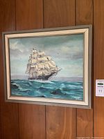 Full view of framed painting on wood-paneled wall showing sailing ship on ocean.