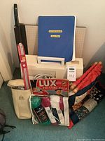 Photo of sewing machine case, cutter board, knitting bag, yarn, needles, and knitting book arranged on floor.