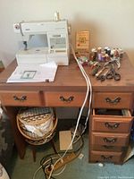 Full view of sewing machine on a wooden sewing desk with drawers and various sewing accessories spread on top including thread spools, scissors, pliers, and an instruction manual.