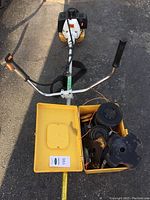 Top-down view showing brush cutter handlebar, yellow case with extra blades and trimmer heads inside the case.