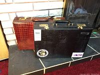 Two vintage-style briefcases side by side on floor near brick fireplace. One is light brown with crocodile pattern, the other dark brown/black with similar textured pattern and stickers.