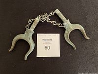 Pair of bronze oar locks laid parallel on a black background with chain linking them, showing an overall view of the lot.