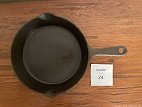 Top view of black cast iron skillet with handle and pouring spouts, on wooden surface with lot number card.