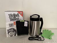 Showcases Magical Butter MB2e stainless steel extractor pitcher with attached black base, green heat-resistant glove, black nylon filter bag, owner’s manual, and power cord laid out on table.