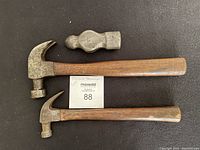 Three Plumb hammer pieces: two claw hammers with wooden handles, one hammer head without handle laid flat on black surface.