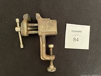 Small metal bench vice shown on black surface with MaxSold lot card, front angled view showing screw adjustment and clamp base.