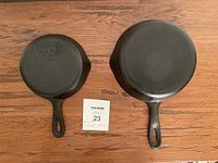 Two Wagner Ware cast iron pans, sizes #3 and #5, shown from the bottom with clear brand markings, placed next to a lot number card on a wooden surface.
