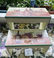 Front view of two-step wooden stool with painted wine bottle, glasses, grapes, cheese, and crates design on steps.