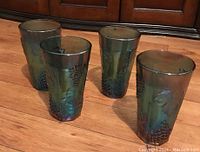 Four carnival glass tumblers arranged on wooden floor showing iridescent Harvest Grape grape cluster and leaves pattern.