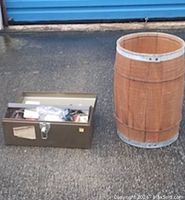 Photo showing a wooden barrel and an open metal toolbox with assorted tools and hardware inside, placed outdoors on pavement.