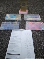 Photo of four sealed plastic containers with assorted beads, an instruction sheet, and a pink plastic stand on a gray surface.