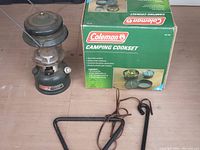 Photo showing boxed Coleman camping cook set and vintage Coleman fuel lantern side by side on floor.