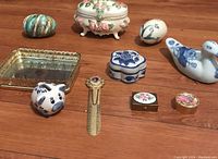 Lot of 10 collectible decorative items on wooden surface, including porcelain trinket boxes, a duck figurine, pig-shaped item, brass bookmark, and decorative mirrored tray.