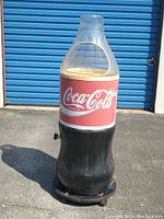 Outdoor photo showing the full Coca-Cola bottle-shaped cooler on wheels with clear plastic dome lid and red Coca-Cola label.