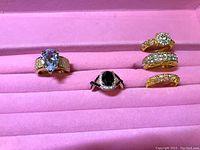 Five rings displayed in a pink velvet ring holder, showing various gemstone styles including purple, black, and clear stones in gold-tone bands.