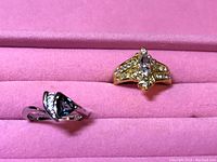 Photo shows two decorative rings on pink velvet background. One gold-toned with purple and clear stones, the other silver-toned with clear stones.