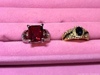 Photo showing two rings placed on pink fabric. One silver-tone with large red square gemstone and clear side stones, one gold-tone with oval dark blue/purple gemstone and clear surrounding stones.