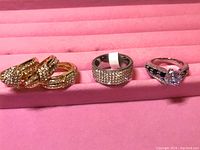 Photo showing the three rings placed on a pink display tray: two gold-tone and silver-tone rings with various crystal and gemstone embellishments.
