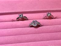 Three solitaire-style rings with clear stones set on a pink velvet display holder.