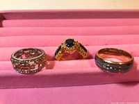 Photo shows three rings laid out on a pink padded display surface. From left to right: silver-tone openwork floral ring, gold-tone ring with dark blue central gemstone and smaller stones, gold-tone band ring with blue inlays.