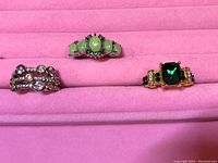 Photo showing three rings displayed on a pink jewelry holder: one with green oval stones, one with clear stones, and one with a large square green stone in gold setting.