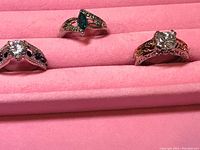 Three rings arranged on pink velvet display; each ring has unique gemstone and intricate metalwork.