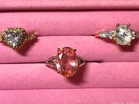 Photo of three rings displayed on a pink cushioned jewelry holder: a heart-shaped clear stone ring, an oval pink stone ring, and a marquise-shaped clear stone ring with smaller clear side stones.