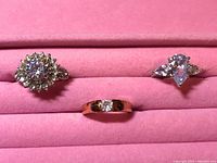 Three gold-tone rings with clear stones displayed on a pink slot jewelry holder