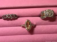 Photo showing three gold-tone rings on a pink soft jewelry holder. Rings include one with red gemstones, one with a marquise-shaped central stone, and one with multiple clear rhinestones arranged in a cluster.