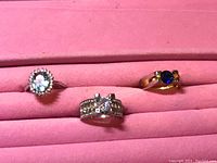Photo of three rings in a pink ring display case: left with light blue oval gemstone and clear stones, center wide band with clear stones, right gold-tone with dark blue stone
