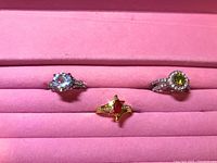 Photo of three rings displayed on a pink velvet tray: silver-tone with clear stones, gold-tone with red stone, and silver-tone with green stone and clear stones.