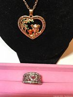 Photo showing a heart-shaped pendant necklace with a dog figure and green stones, and a rhinestone ring with embedded rhinestones on a black stand.
