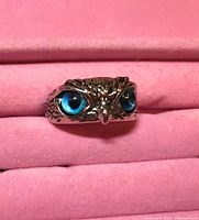 Close-up image of a silver-toned owl head ring with detailed feathers and bright blue eyes, displayed in a pink cushioned tray.