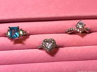 Photo showing three rings in a pink padded display holder: one with a blue rectangular gemstone and side stones, one with a round center gemstone and small stones on the band, and one with a heart-shaped center stone surrounded by small stones.