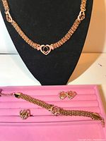 Photo of a 5-piece gold-tone jewelry set displayed against black and pink backgrounds showing necklace on bust, bracelet, ring, and earrings