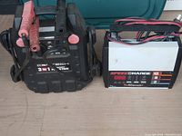 Photo showing a black Ceratech 3 in 1 portable jump starter pack with attached jumper cables and a white SpeedCharge 12 Amp battery charger with cables placed on a wooden surface.