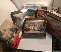 Photo of puzzle boxes arranged showing one 3000 piece Ravensburger box and multiple 1000 piece puzzles including two Thomas Kinkade puzzles, a Hershey's Masterpieces puzzle, and a 500 piece Castle puzzle.