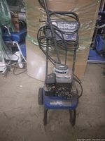 Front view of a Campbell Hausfeld pressure washer, showing the metal frame, wheels, motor, pump, power cord neatly coiled on the handle, and hose with spray wand attached.
