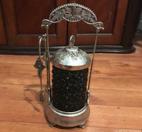 Front view of the silver plated Victorian style pickle castor with black pressed glass jar showing ornate floral design on the arched handle and lid.