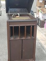 Photo of the entire vintage record player cabinet showing the open lid with the tonearm and turntable visible. The front shows speaker grates and storage doors below.