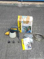 Photo showing Wagner Power Painter system components including spray gun with canister, flexible hose, two nozzles, user manual, and product box placed on pavement.