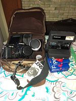 Full view of Chinon CG-5 camera with attached lens inside brown leather case, alongside Polaroid camera and film boxes.