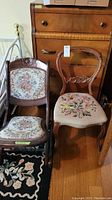 Two vintage chairs side by side in front of wooden furniture; one with rounded back and floral needlepoint seat, the other a wooden folding rocker with floral needlepoint upholstery