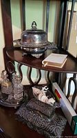 Photo showing the wooden onyx scale with elephant weights, crystal decanter cruet set with silver lids on a silver holding tray, and a glass bottle on shells along with a small Bible book on the shelf.