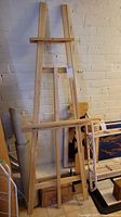 Overall view of the wooden artists easel leaning against a white brick wall, showing the full structure and canvas holder.