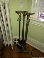 Full view of the brass fireplace tool set standing by a radiator, showing four tools with horse head handles and the brass stand.